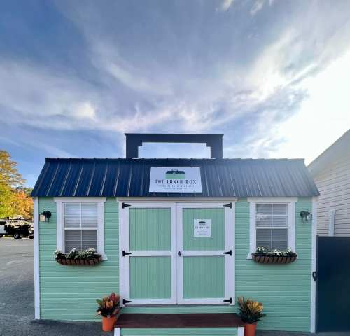 A small, green food stand with a blue roof, labeled "The Lunch Box," surrounded by plants and a clear sky.