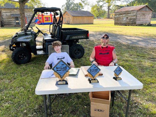 Two men sit at a table with trophies, outdoors near a utility vehicle, in a park-like setting.