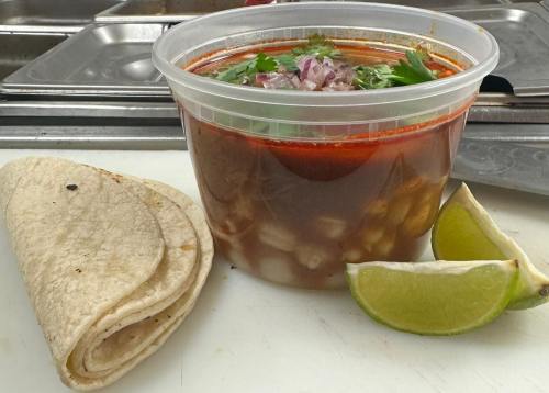 A bowl of rich red soup topped with onions and cilantro, alongside two tortillas and lime wedges on a cutting board.
