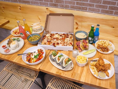 A table filled with various dishes: pizza, sandwiches, salads, fried chicken, fries, and colorful drinks.
