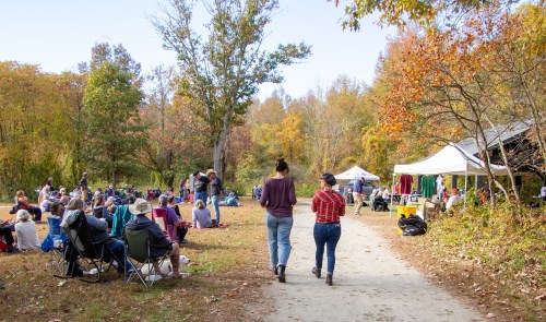 Two women walk along a path in a park, surrounded by people enjoying an outdoor event amid autumn foliage.