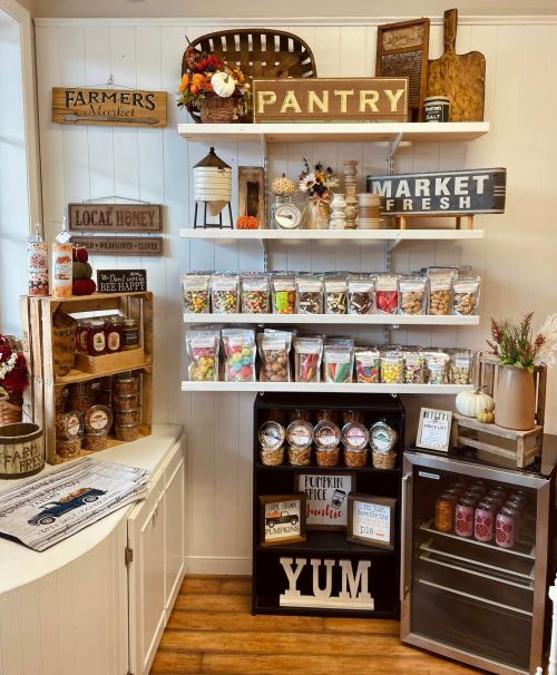 A cozy pantry display with shelves of snacks, jars, and signs, featuring a mini fridge and autumn decorations.