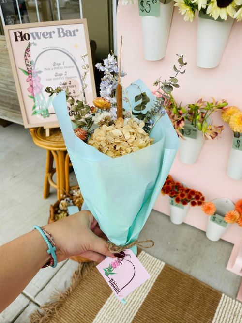 A hand holds a pastel blue bouquet with dried flowers, in front of a flower bar display and a sign.