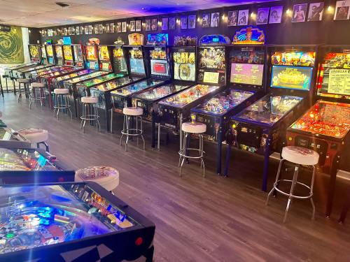 A vibrant arcade featuring rows of colorful pinball machines and bar stools, with framed photos on the wall.