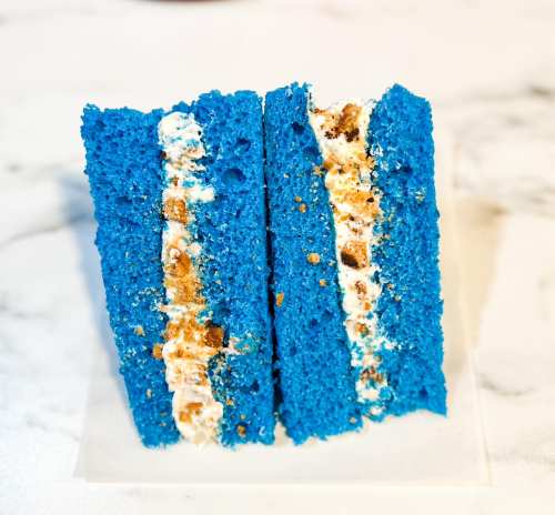 A vibrant blue cake slice with a creamy filling and crumbled topping, displayed on a white surface.