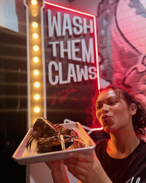 A person holds a tray of seafood in front of a neon sign that says "WASH THEM CLAWS."