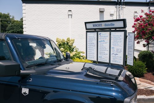 A car parked in front of a restaurant menu board at Biscuit Doodle, with flowers and greenery in the background.