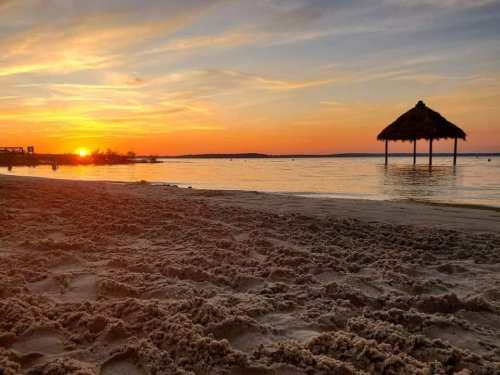 Sunset over a calm lake, with a sandy beach in the foreground and a thatched gazebo on the water's edge.