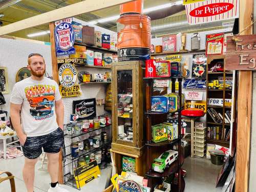 A man stands in an antique shop surrounded by vintage signs, collectibles, and various nostalgic items.