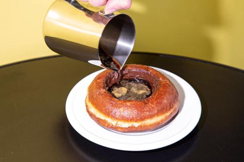A hand pours chocolate sauce into a round pastry on a black table, with a yellow background.