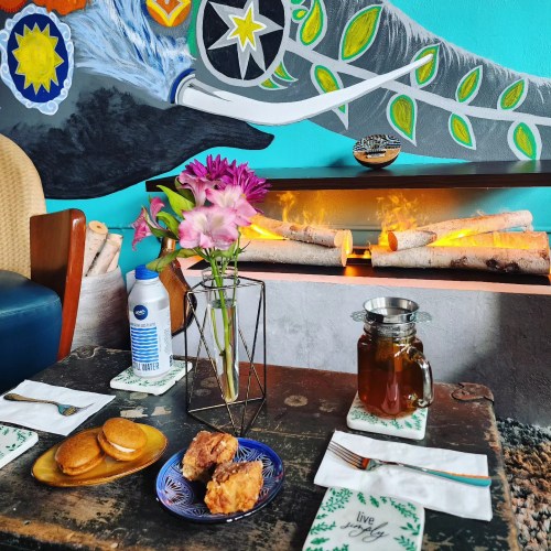 A cozy table setting with a vase of flowers, snacks, and a drink, against a colorful mural and a glowing fireplace.