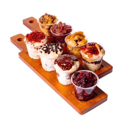 A wooden tray holds eight small dessert cups filled with various toppings like chocolate, fruit, and nuts.