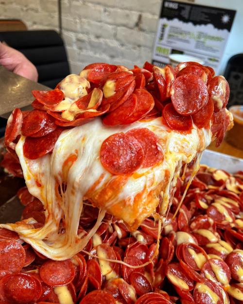A slice of cheesy pizza topped with pepperoni, being lifted from a tray overflowing with more pepperoni.
