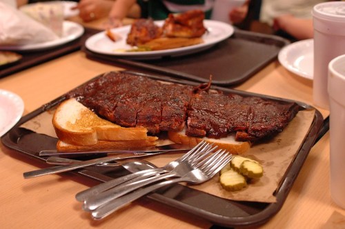 A plate of barbecue ribs with slices of bread and pickles, served on a tray with utensils.