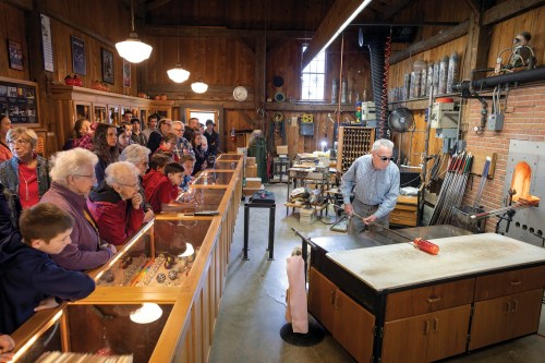 A glassblowing demonstration in a workshop, with a crowd watching as an artist shapes molten glass.