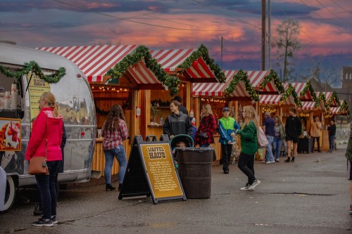 A festive market with striped tents, food trucks, and people enjoying the evening under a colorful sky.