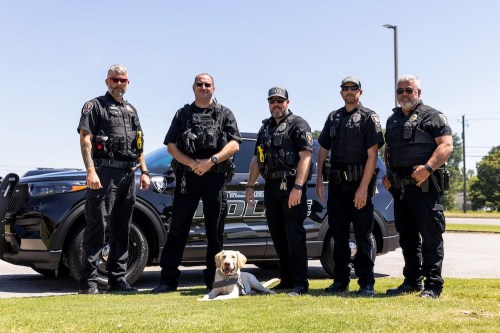 Five police officers in tactical gear stand beside a police vehicle, with a yellow labrador sitting in front of them.