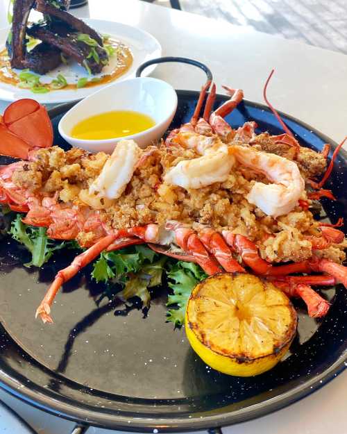 A beautifully presented lobster dish topped with seafood stuffing, served with grilled lemon and olive oil on a black plate.