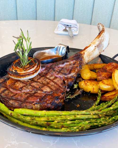 A large grilled tomahawk steak garnished with rosemary, served with roasted potatoes, asparagus, and a small sauce dish.