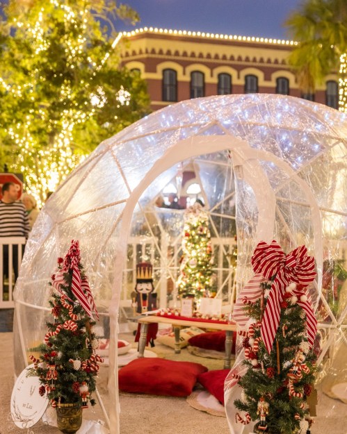 A festive igloo with Christmas trees, red pillows, and twinkling lights, surrounded by holiday decorations.