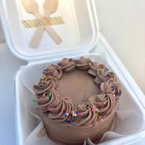 A small chocolate cake with swirled frosting and colorful sprinkles, placed in a takeout container.