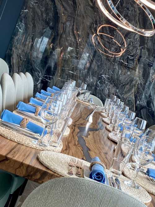 Elegant dining setup with a long wooden table, blue napkins, and glassware against a marble backdrop.