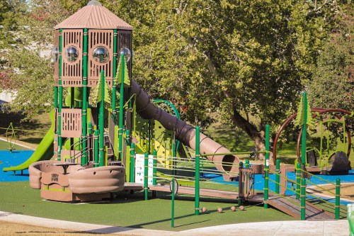 Colorful playground featuring a large slide, climbing structures, and swings, surrounded by green trees and grass.