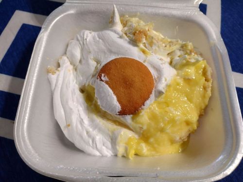A takeout container with banana pudding topped with whipped cream and a vanilla wafer.