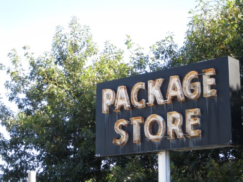 A weathered sign reading "PACKAGE STORE" against a backdrop of green trees and a clear blue sky.