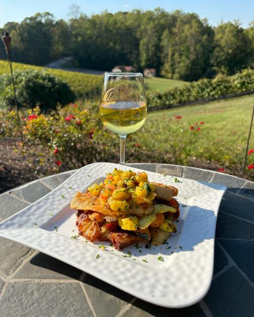 A plate of grilled chicken with pineapple salsa, accompanied by a glass of white wine, set against a scenic vineyard backdrop.