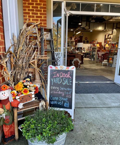 A sign outside a store advertises an indoor yard sale every weekend, with fall decorations and a welcoming entrance.