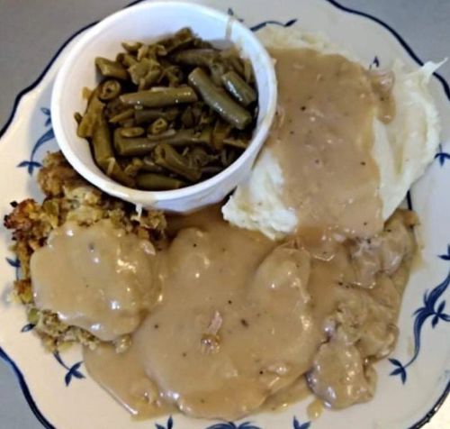 A plate of comfort food featuring mashed potatoes with gravy, green beans, stuffing, and chicken with gravy.