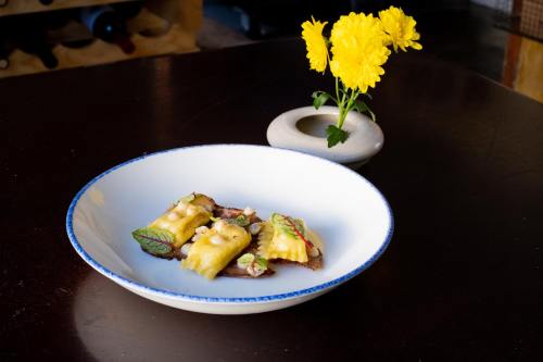 A white bowl with colorful ravioli on a dark table, accompanied by a small vase of yellow flowers.