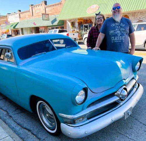 A couple stands beside a vintage blue car on a sunny street with shops in the background.