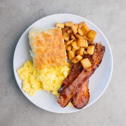 A plate of scrambled eggs, crispy bacon, golden breakfast potatoes, and a biscuit.