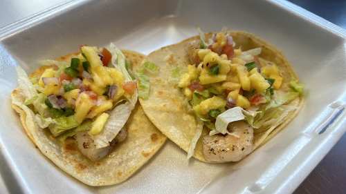 Two shrimp tacos topped with lettuce, mango salsa, and diced tomatoes in a takeout container.