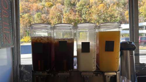 Four beverage dispensers filled with iced tea, lemonade, and other drinks, set against a backdrop of autumn foliage.