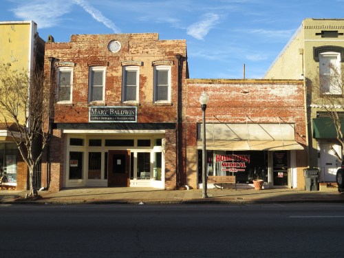 Historic brick buildings with storefronts, including "Mary Baldwin" and "Hometown Medical," under a blue sky.
