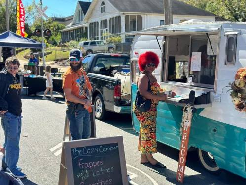 A food truck with a sign reading "The Cozy Camper" serves customers at an outdoor event on a sunny day.