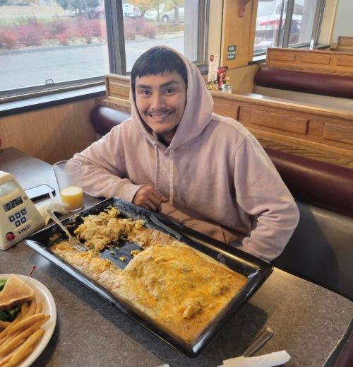 A smiling person in a hoodie sits at a diner table with a large plate of food, including a cheesy omelet and rice.