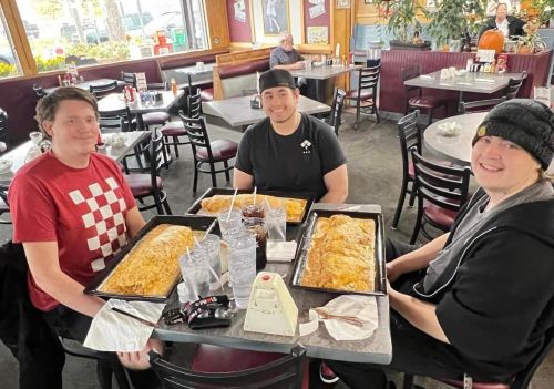 Three friends sit at a restaurant table with large trays of food, smiling and enjoying their meal together.