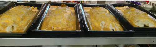 Four large trays of enchiladas covered in cheese and sauce, displayed on a countertop in a restaurant kitchen.
