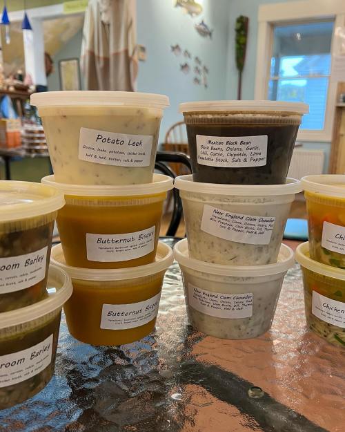 A variety of labeled soup containers stacked on a table, including flavors like Potato Leek and New England Clam Chowder.