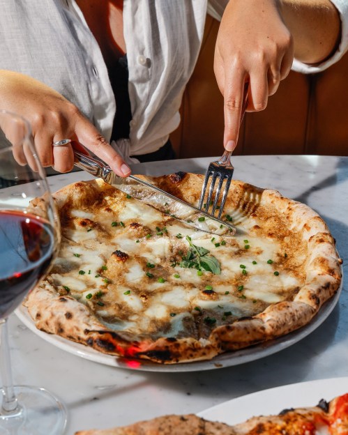 A person cuts into a cheese pizza topped with herbs, with a glass of red wine nearby on a marble table.