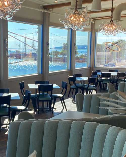 Bright restaurant interior with blue chairs, large windows overlooking a waterfront, and decorative light fixtures.