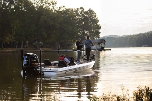 Two people on a fishing boat in calm waters, with trees and a dock in the background under a soft morning light.