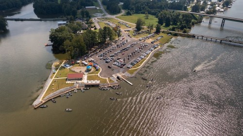 Aerial view of a lakeside area with a parking lot, boats, and green spaces, surrounded by water and trees.