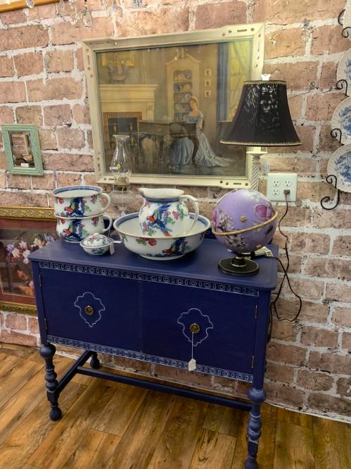 A vintage blue cabinet with decorative porcelain bowls, a lamp, and a globe, set against a brick wall and framed artwork.