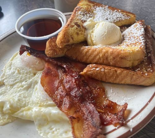 A plate of French toast topped with powdered sugar and ice cream, served with bacon and eggs, alongside syrup.