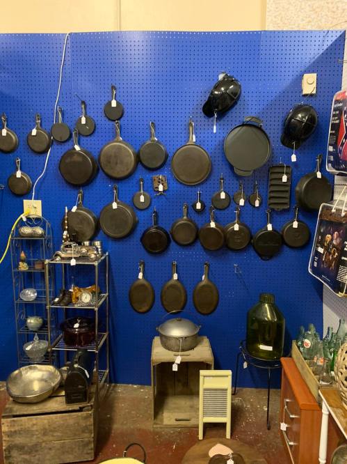 A display of various cast iron pans and cookware hanging on a blue pegboard wall, with additional items on shelves below.